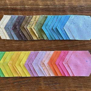 May include: Two rows of colorful fabric pieces in a chevron pattern. The top row transitions from white to blue, while the bottom row goes from green to pink. Each piece has a hexagonal shape with a speckled design.