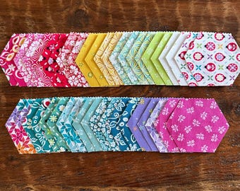 May include: A collection of fabric hexagons arranged in two rows, featuring a variety of colors including pink, red, yellow, green, teal, blue, and purple. Each piece displays distinct floral, geometric, and ladybug patterns with pinked edges.