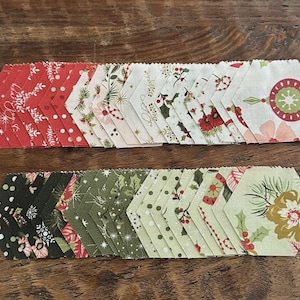 May include: A collection of fabric hexagons arranged in two rows. The top row features red and cream fabrics with holiday-themed prints including snowflakes, pine needles, and ornaments. The bottom row displays black, dark green, and light green fabrics.