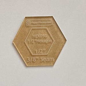 May include: A clear, tan-colored acrylic hexagon quilting template. The template has the text "PaperPieces.com", "HEX050", "1/2" Hexagon", "1/2"", and "3/8" Seam" etched into the surface. The hexagon measures 1/2 inch across.