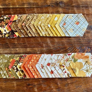 May include: A collection of fabric hexagons arranged in two horizontal rows. The fabric pieces feature various patterns including floral prints, plaid, and small pumpkin motifs in a color palette of orange, yellow, brown, green, and cream.