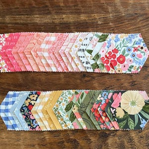 May include: A collection of fabric hexagons arranged in two horizontal rows. The top row features pink, coral, and white floral and gingham patterns. The bottom row displays blue, yellow, green, and dark floral prints with various botanical designs.
