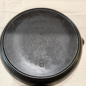 May include: A black cast iron skillet with the text "WAGNER SIDNEY" and the number "8" embossed on the bottom. The skillet has a round cooking surface and a handle.