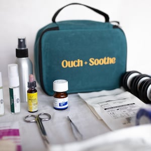 Könnte beinhalten: Ein türkisfarbener Erste-Hilfe-Kasten mit den Worten "Ouch + Soothe" in goldfarbenen Buchstaben. Der Kasten ist von verschiedenen medizinischen Hilfsmitteln umgeben, darunter Flaschen, Gläser, Scheren und Bandagen. Für kleinere Verletzungen.
