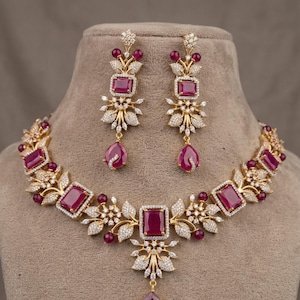 Puede incluir: Un conjunto de joyas de tono dorado con un collar y pendientes a juego. El collar y los pendientes están adornados con grandes piedras preciosas de color rojo rubí rectangulares, rodeadas de piedras claras más pequeñas y detalles florales. Los pendientes tienen un colgante de rubí en forma de lágrima.