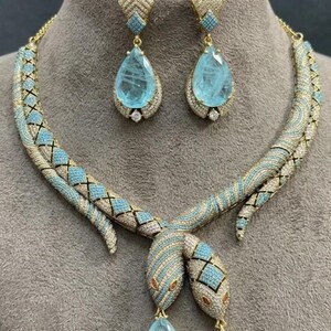 May include: A jewellery set featuring a necklace and matching drop earrings. The necklace is designed as two intertwined snakes with gold-tone metal, pavé-set clear crystals, and light blue accents, each ending in a large, pear-shaped light blue stone.