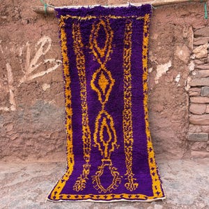 May include: A rectangular, purple rug with a yellow geometric pattern. The rug is hanging against a textured, brown wall. The rug's design features diamond and floral shapes.