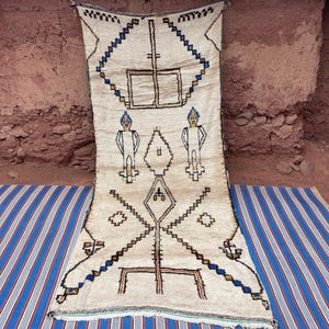 May include: Cream-colored, hand-woven rug with geometric patterns in black, brown, and blue. The rug features diamond, rectangular, and linear designs, with two stylized human figures. The rug is displayed on a blue and white striped surface.