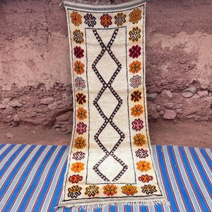 May include: A rectangular, cream-colored rug with a central diamond pattern in dark brown. The rug is bordered by repeating floral motifs in shades of orange, yellow, and red. The rug is displayed on a blue and white striped surface.