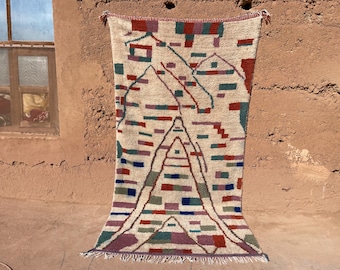 May include: A rectangular, cream-colored rug with a geometric design. The rug features a central triangle and mountain-like shapes, with accents in red, blue, green, and purple. The rug has a fringe at the bottom and is hanging against a textured wall.