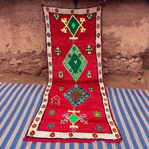 May include: A vibrant red rug with geometric designs in green, yellow, and blue. The rug features a white border with floral accents. The rug is placed on a blue and white striped surface.