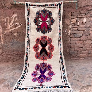 May include: Cream-colored rectangular rug with three floral designs in the center. The floral designs feature red, black, gray, and purple accents. The rug has a black border with blue and red details and a fringed edge.