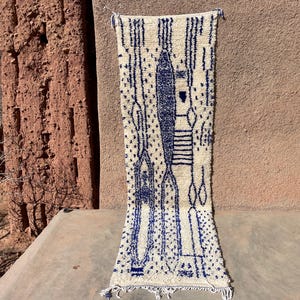 May include: A rectangular, off-white rug with a blue geometric design. The design includes vertical lines, dotted patterns, and stylized shapes. The rug is hanging against a textured, reddish-brown wall.