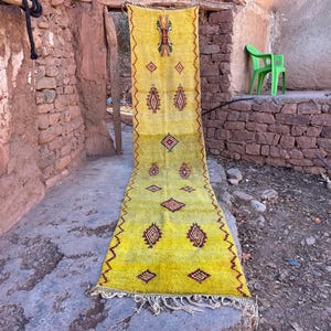May include: A long, rectangular yellow rug with a geometric pattern. The rug has a central design of diamonds and other shapes in red and brown. The rug is hanging in an outdoor setting, with a stone wall and a green chair in the background.