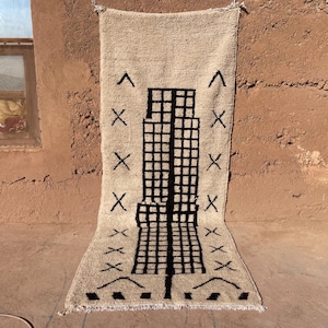 May include: Cream-colored, rectangular rug with a geometric design. The rug features a central black grid pattern, flanked by black 'X' shapes and arrow-like symbols. The rug is hanging against a textured, reddish-brown wall.