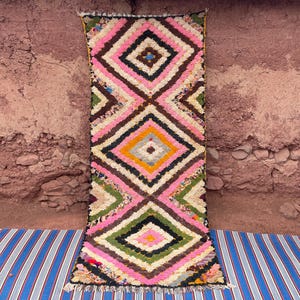 May include: A rectangular, colorful rug with a diamond pattern. The rug features pink, white, brown, green, and yellow geometric shapes. The rug is displayed on a blue and white striped mat against a reddish-brown wall.