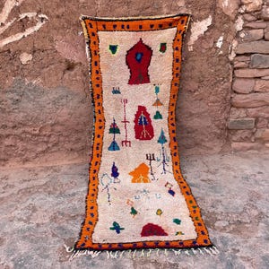 May include: A rectangular, cream-colored rug with an orange border and black trim. The rug features colorful geometric shapes and symbols in red, blue, green, and orange. The rug is displayed against a textured, reddish-brown wall.