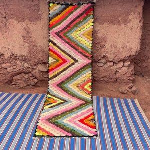 May include: A colorful, rectangular rug with a zig-zag pattern in shades of pink, yellow, red, green, and brown. The rug is placed on a blue and white striped mat, with a textured, reddish-brown wall in the background.