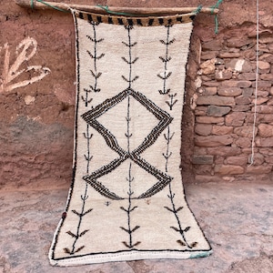 May include: A cream-colored, rectangular rug with a geometric design. The rug features a central diamond shape and vertical lines with arrow-like details. The rug is hanging against a textured, reddish-brown wall.