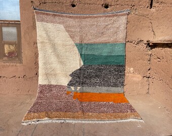 May include: A rectangular, geometric patterned rug with a mix of colors including beige, brown, teal, black, orange, and burgundy. The rug is hanging against a textured, reddish-brown wall. The design features a large, abstract shape.