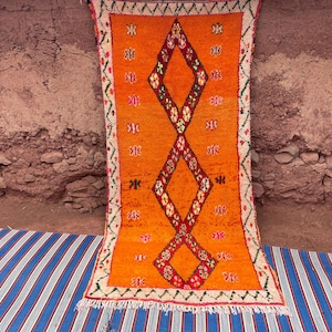 May include: An orange Moroccan rug with a diamond pattern in red and white. The rug has a cream-colored border with green and red accents and a fringed edge. The rug is displayed against a textured brown wall and a blue and white striped surface.
