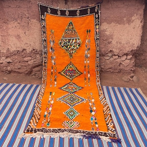May include: An orange Moroccan rug with geometric diamond and triangle patterns. The rug has a dark brown border and colorful accents. It is displayed on a blue and white striped mat against a textured wall.