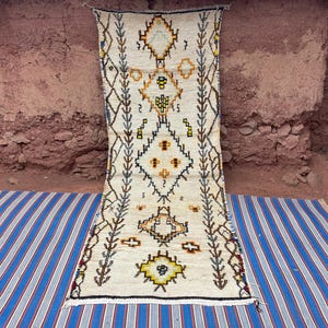 May include: Cream-colored, rectangular rug with geometric patterns in yellow, orange, and black. The rug features diamond and cross motifs, with a border of stylized plant-like designs. It is displayed on a blue and white striped surface.