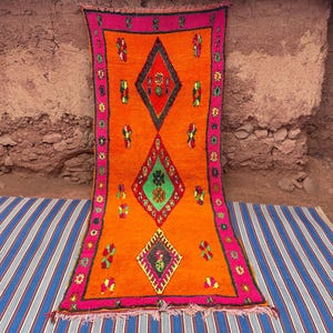 May include: An orange rug with a geometric diamond pattern in red, green, and black. The rug has a pink border with floral accents. The rug is displayed on a blue and white striped mat.