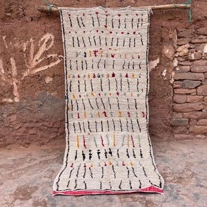 May include: A rectangular, off-white rug with a geometric pattern of vertical black lines and colorful accents. The rug is hanging against a textured, reddish-brown wall. The bottom edge has a pink border.