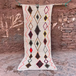 May include: A beige rug with a diamond pattern in brown, black, yellow, red, and white. The rug is hanging from a wooden rod against a textured, reddish-brown wall. The rug's design features alternating diamond shapes.