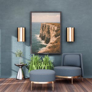 May include: A coastal-themed interior design featuring a framed landscape photograph of a cliffside overlooking the ocean. The room includes a blue armchair and ottoman with gold trim, two wall sconces, and a small silver side table with greenery.