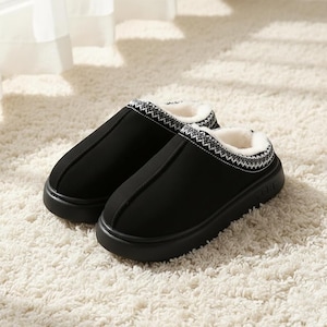 Puede incluir: Un par de zapatillas negras con suela gruesa e interior suave y blanco. El borde superior presenta un patrón decorativo en blanco y gris. Las zapatillas están diseñadas para uso en interiores y están colocadas sobre una alfombra blanca.