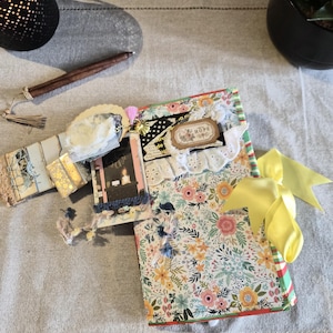 Può includere: Un diario rettangolare con un motivo floreale di fiori rosa, gialli e blu. La copertina è decorata con pizzo, una piccola etichetta con la scritta HOPE, un fiocco giallo e un ciondolo con un'illustrazione in miniatura di una persona.