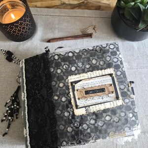 May include: A square journal with a black lace spine and a cover featuring a gray circular pattern overlay. A central rectangular label displays the word JOURNAL, accented with wire mesh, metal brads, and a background of vintage French text.