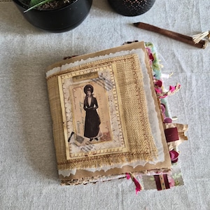 May include: A handmade journal with a burlap cover featuring a vintage-style postcard image of a person in a dark coat and hat. The cover includes the text NEW MEMORIES and CARTE POSTALE. The edges are layered with textured paper and fabric scraps.