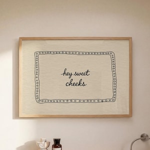 Puede incluir: Una impresión enmarcada con el texto "hey sweet cheeks" en un baño. Debajo, una estantería de madera con objetos decorativos y una botella marrón oscuro. Un lavabo con dispensador de jabón, grifo y portacepillos.