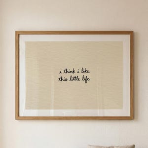May include: A framed print with the text "i think i like this little life" in a black script font. The print has a beige background and is set in a light brown wooden frame. The print is hanging above a beige sofa with a pillow.