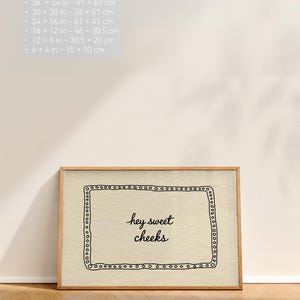 May include: A framed print with the text "hey sweet cheeks" in a handwritten style. The print is in a wooden frame and has a decorative border. The image also includes size options in inches and centimeters.