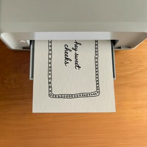 May include: A sheet of paper with the words "hey sweet cheeks" printed in black cursive font is emerging from a gray printer. The paper has a black dotted border. The printer sits on a wooden surface.