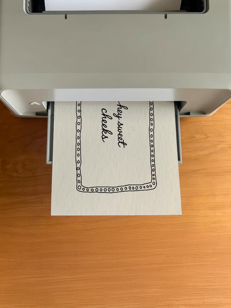 May include: A sheet of paper with the words "hey sweet cheeks" printed in black cursive font is emerging from a gray printer. The paper has a black dotted border. The printer sits on a wooden surface.