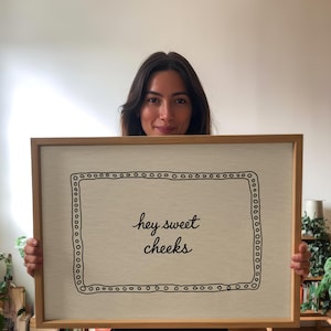 May include: A framed artwork with the text "hey sweet cheeks" in a handwritten style. The artwork has a light beige background with a black dotted border and is encased in a light brown wooden frame.