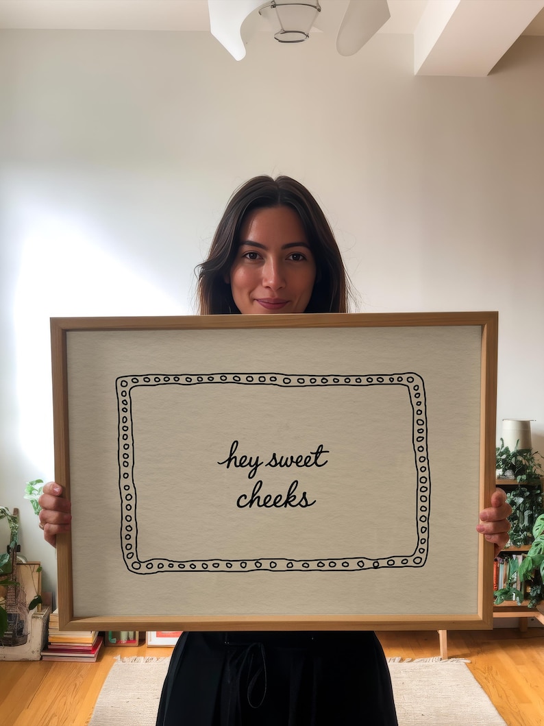 May include: A framed artwork with the text "hey sweet cheeks" in a handwritten style. The artwork has a light beige background with a black dotted border and is encased in a light brown wooden frame.