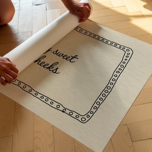 May include: A light-colored rectangular print with the words "sweet cheeks" in cursive, framed by a black dotted border. The print is being unrolled, revealing the design. The background is a light wood floor.