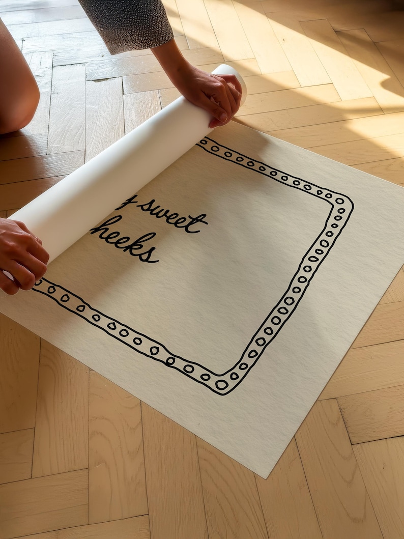 May include: A light-colored rectangular print with the words "sweet cheeks" in cursive, framed by a black dotted border. The print is being unrolled, revealing the design. The background is a light wood floor.