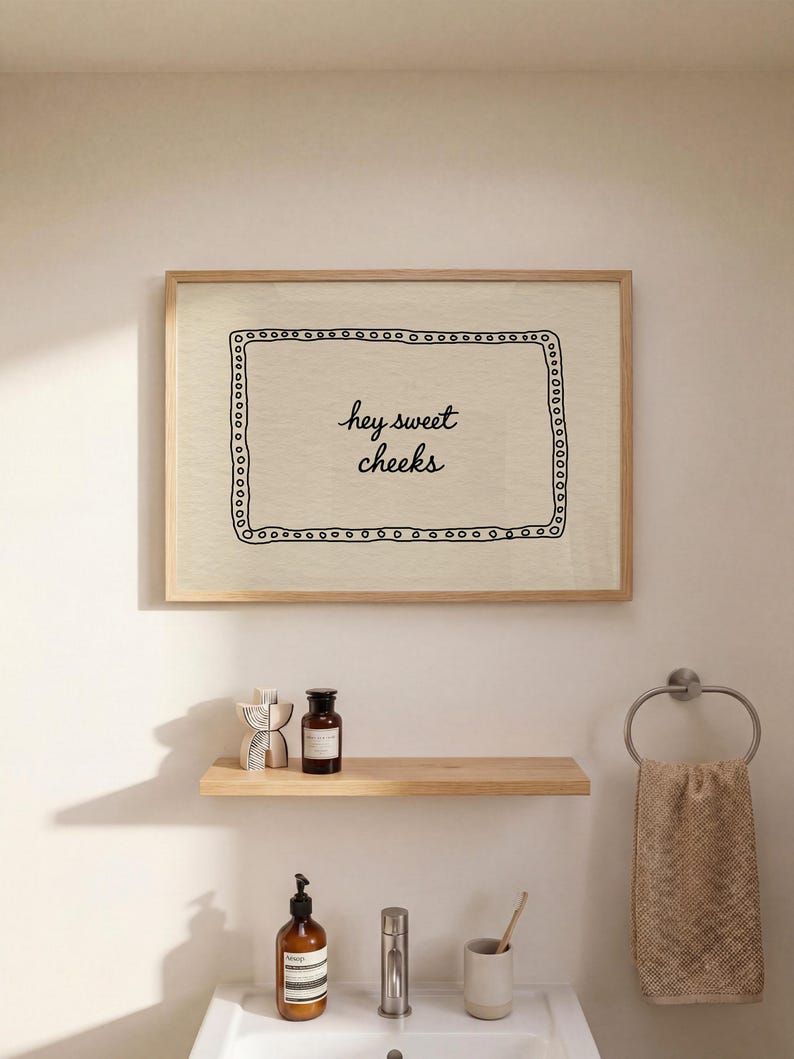 May include: A framed print with the text "hey sweet cheeks" in a bathroom setting. The print is above a wooden shelf holding decorative items and a dark brown bottle. Below, a sink with a soap dispenser, faucet, and toothbrush holder.