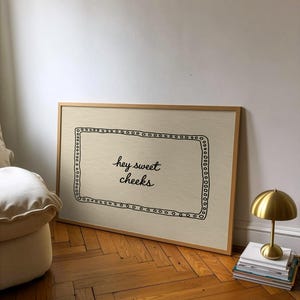 May include: A framed art print with the text "hey sweet cheeks" in black script. The print has a light beige background and a decorative black border. A small gold lamp sits on a stack of books next to the print.