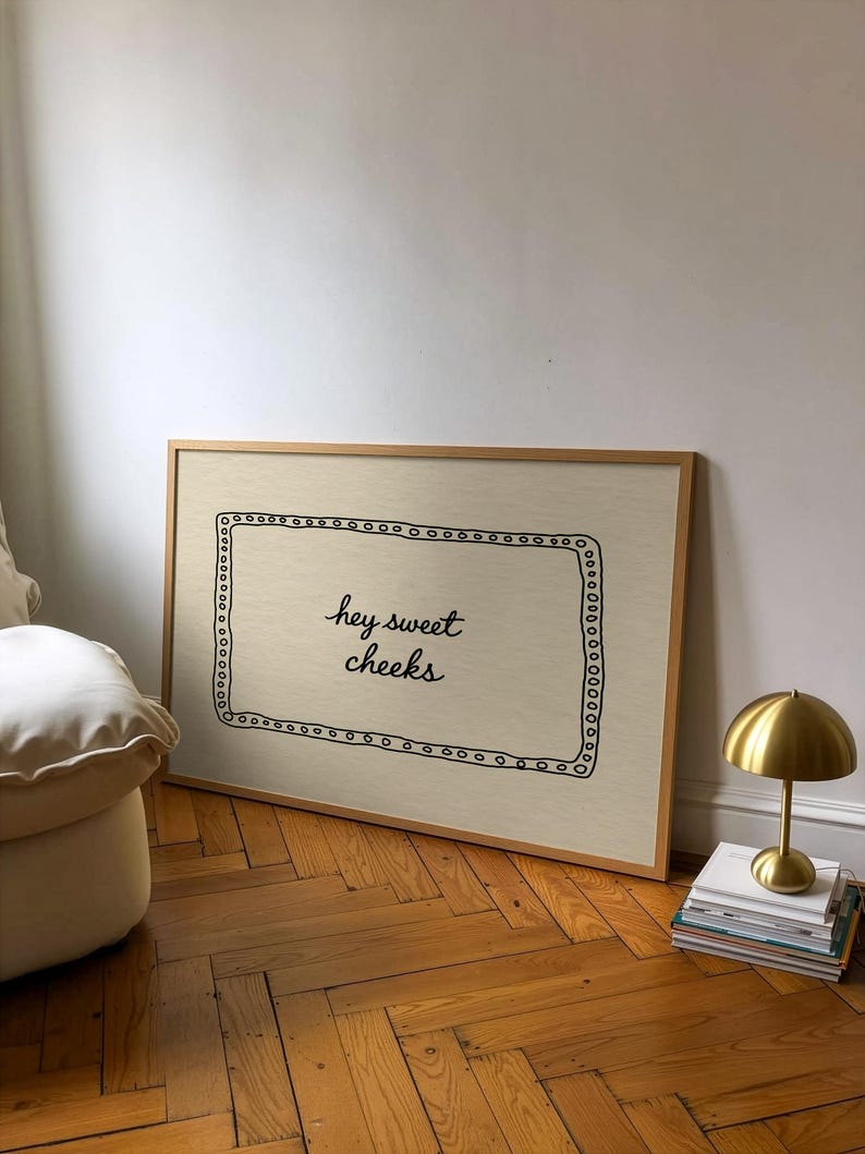 May include: A framed art print with the text "hey sweet cheeks" in black script. The print has a light beige background and a decorative black border. A small gold lamp sits on a stack of books next to the print.