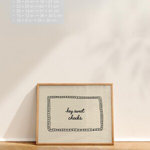 May include: A framed print with the text "hey sweet cheeks" in a handwritten style. The print is in a wooden frame and has a beige background. The image also includes size conversions in inches and centimeters.