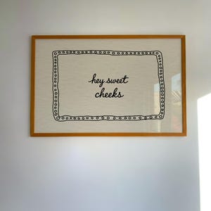 May include: A framed artwork with a light beige background and the phrase "hey sweet cheeks" in black script. The artwork is enclosed in a wooden frame with a decorative border of small black circles.