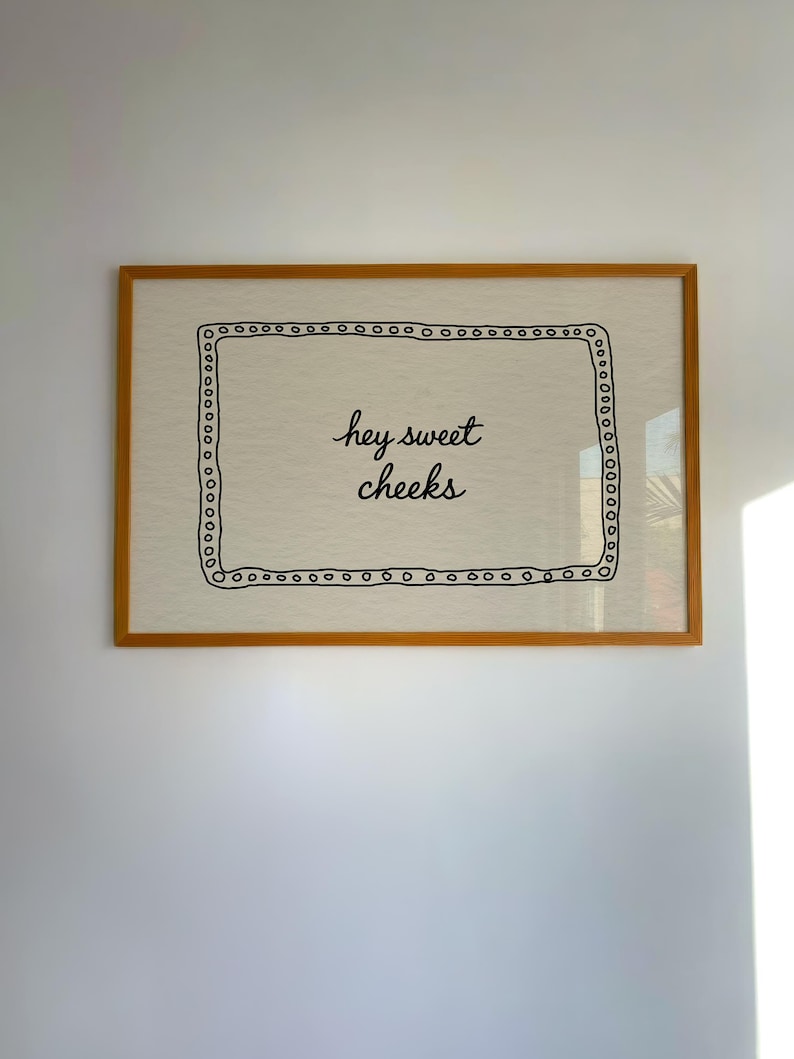 May include: A framed artwork with a light beige background and the phrase "hey sweet cheeks" in black script. The artwork is enclosed in a wooden frame with a decorative border of small black circles.